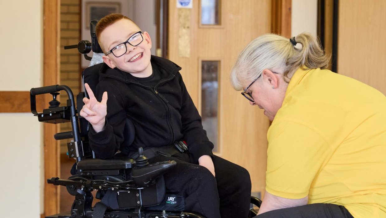 Logan gets his wheelchair adjusted by Senior Mobility Therapist Sarah Wallace in our Billingshurst clinic Logan in his wheelchair, posing for the camera. He's holding his hands up and pointing with his thumbs and fingers. Sarah Wallace is adjusting his wheelchair.