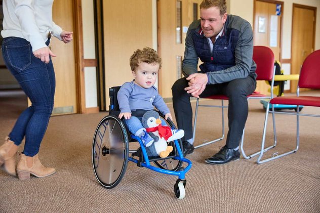 Jake in his wheelchair looking at the camera. He has his toy penguin on the footplate. He is wearing blue Adidas trainers.