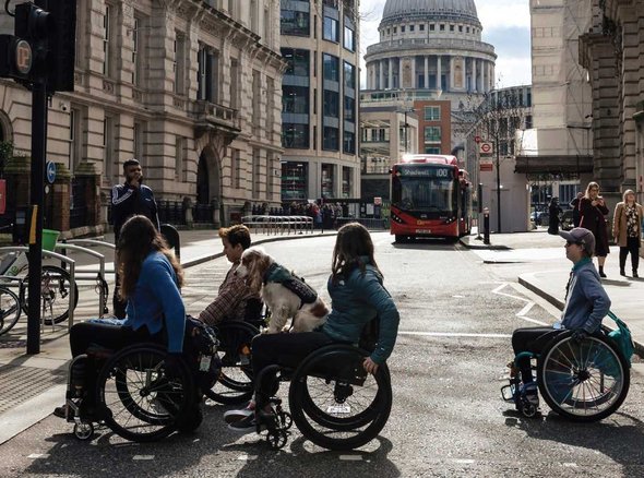Four young wheelchair users cross a Central London street with a single decker bus in the background