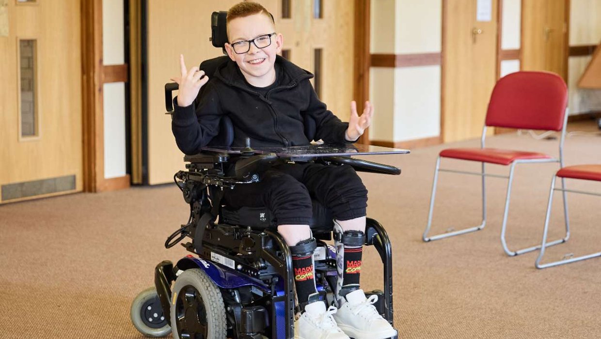 Logan in his wheelchair, posing for the camera. He's holding his hands up and pointing with his thumbs and fingers. He's inside in the clinic, on a beige carpet with a wooden door in the background.