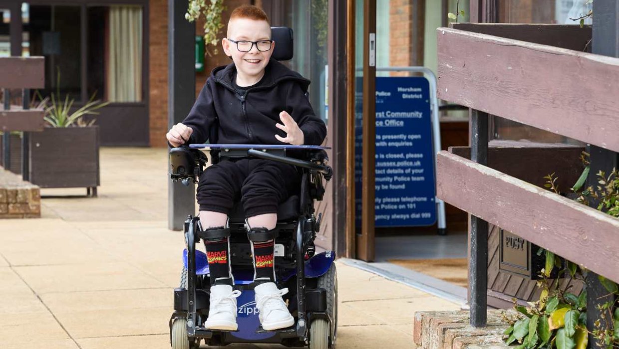 Logan in his wheelchair, outside. He's smiling at the camera. He's outside and wearing black clothes, white trainers and socks with the Marvel Comics logo on.